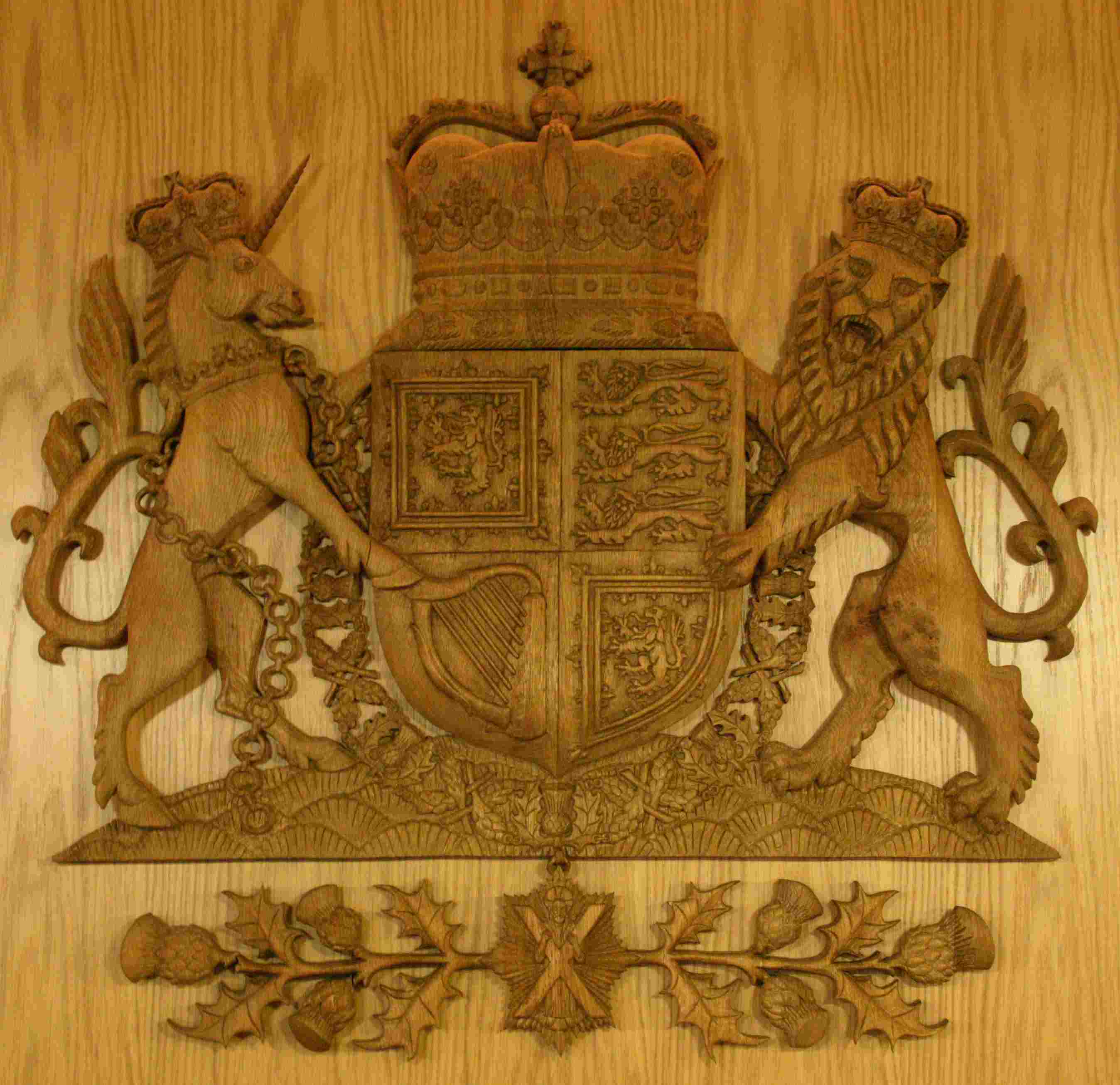 Carved oak coat of arms. Unicorn on the left and lion on the right hold a shield with four quarters. The top left and lower right quarters show the lion rampant flat, the lower left has a harp and the upper right three lions. The shield is topped by a crown. Around the shield is a wreath and underneath is St Andrews holding his cross and a thistle and leaf decoration