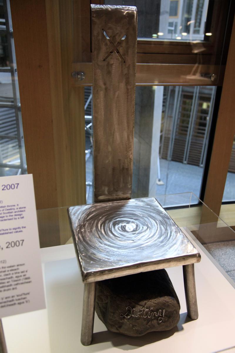 Steel sculpture of a low-seated chair with a cut-out saltire flag in the narrow back of the chair. Under the chair is a boulder with wire on its front in the shape of the word ‘Destiny’.