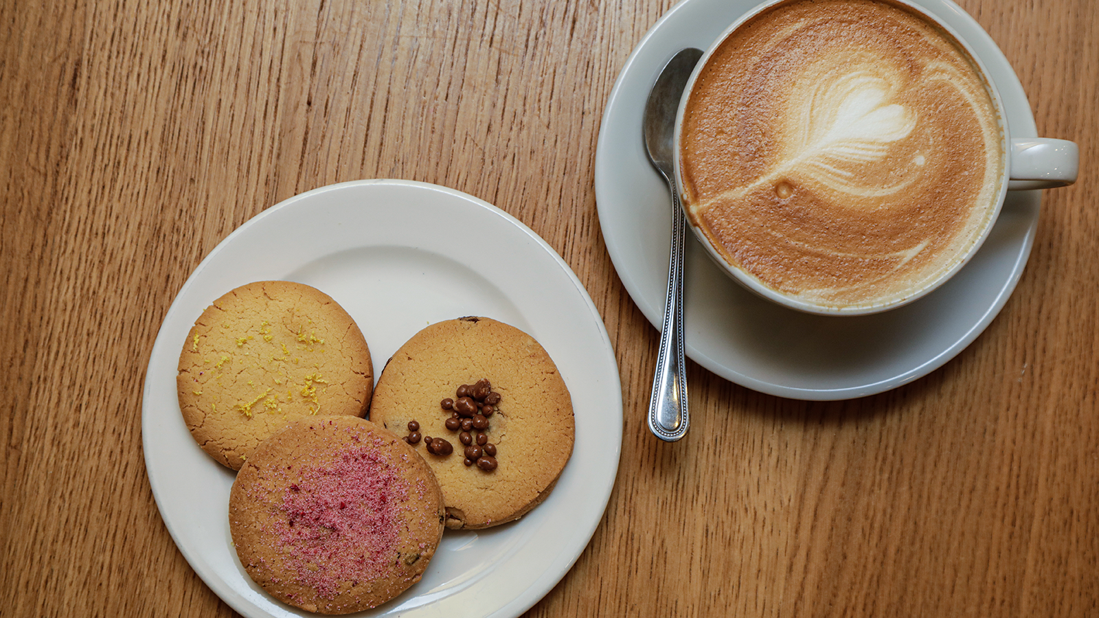 Cappuccino with heart-shaped latte art next to a white plate holding three assorted shortbread biscuits on a wooden table