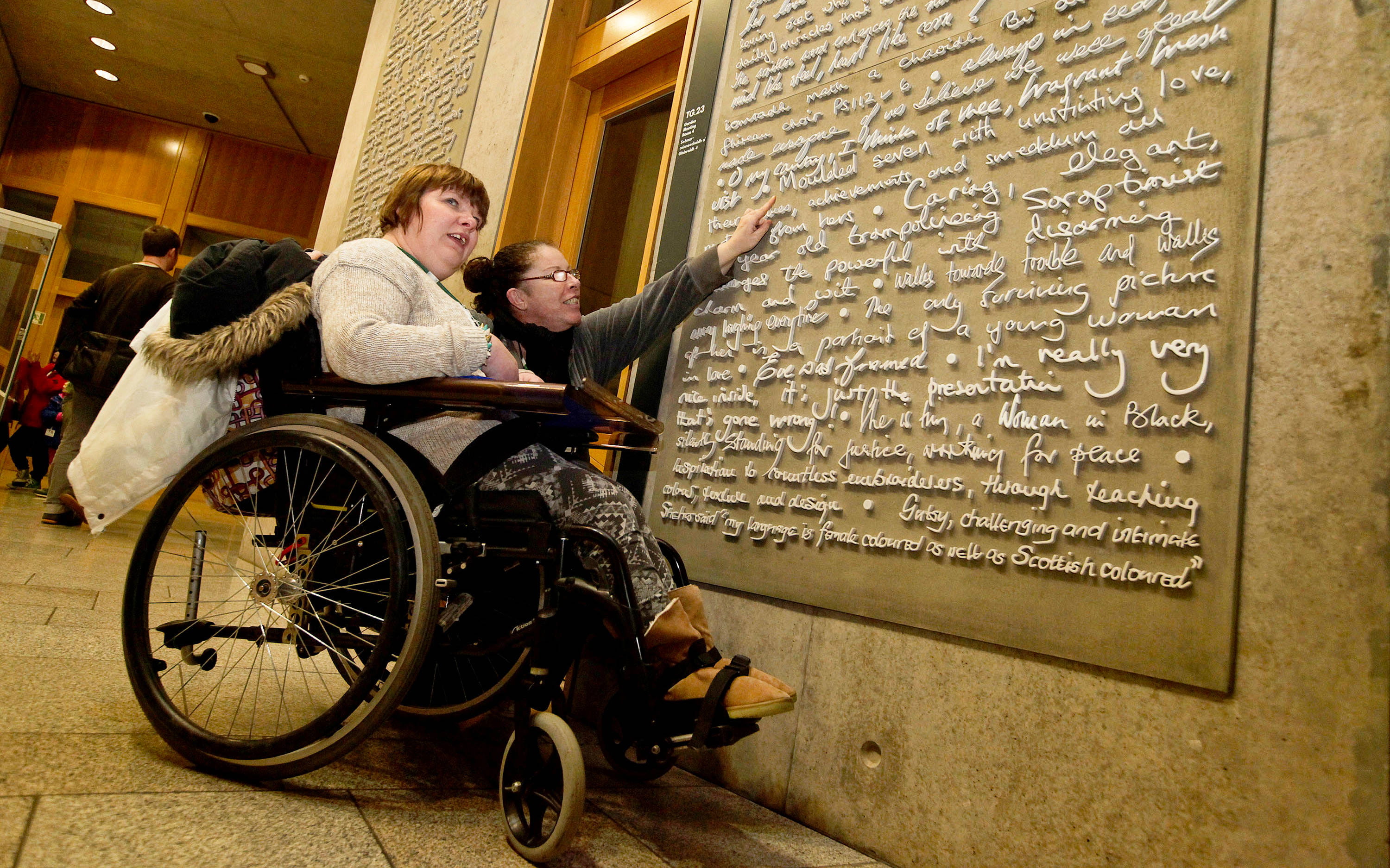 A visitor in a wheelchair and another visitor crouching next to them stop in front of an artwork made up of different handwriting styles displayed on a concrete wall