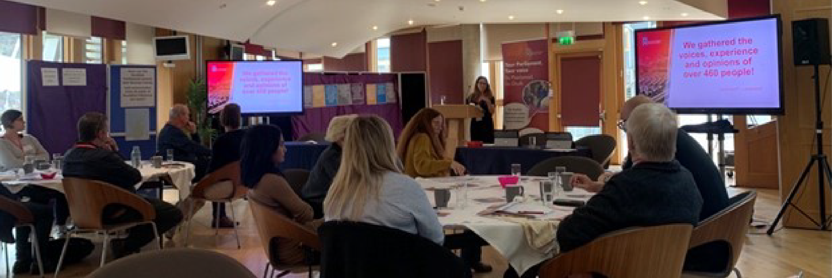 Photo of Citizens' Panel sitting around tables listening to evidence from a witness. The witness is speaking from a lectern in the middle of the room and is standing between 2 large screens that are displaying slides. The slide in the photo reads "We gathered the voices, experiences, and opinions of over 460 people!"